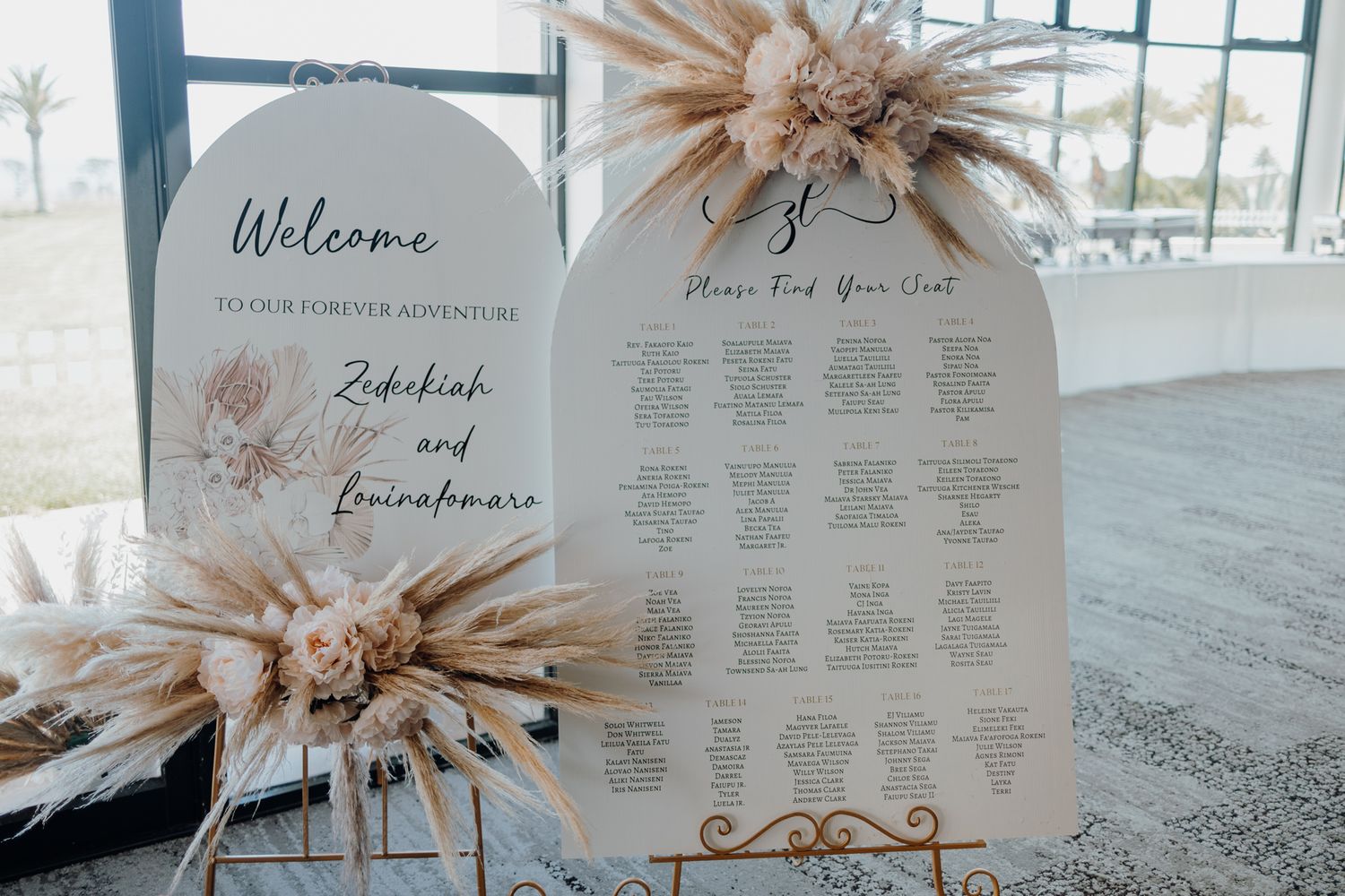 Elegant wedding seating chart signage decorated with dried pampas grass arrangements against a blue wall.