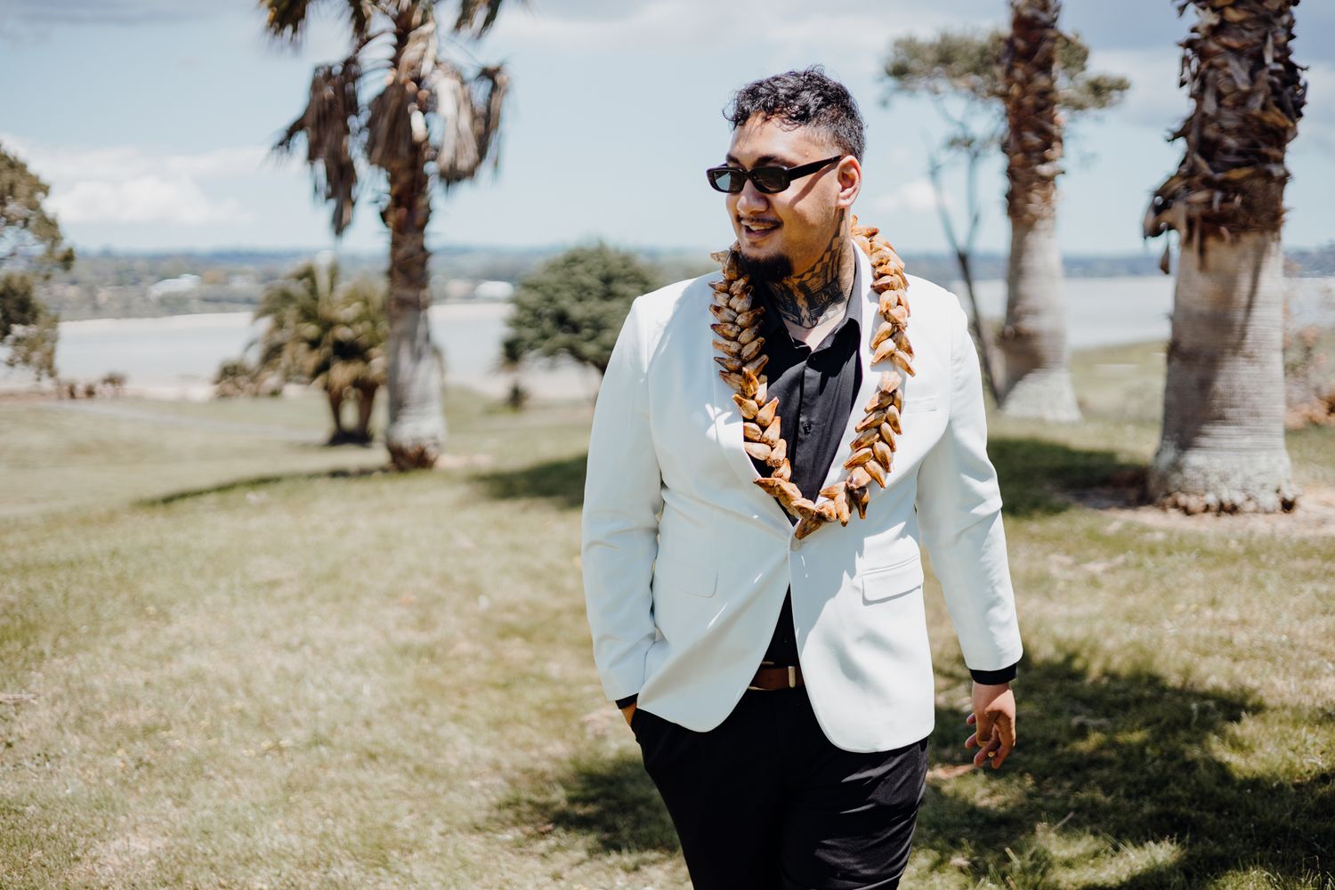 Person in white blazer and lei stands against backdrop of palm trees and coastal scenery.