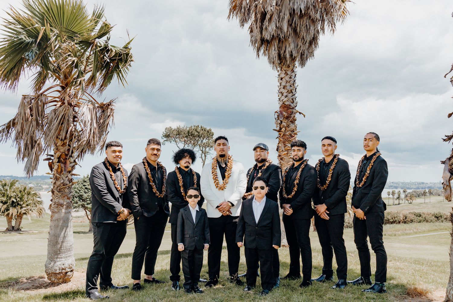 Formal group portrait of wedding party members wearing leis under palm trees.