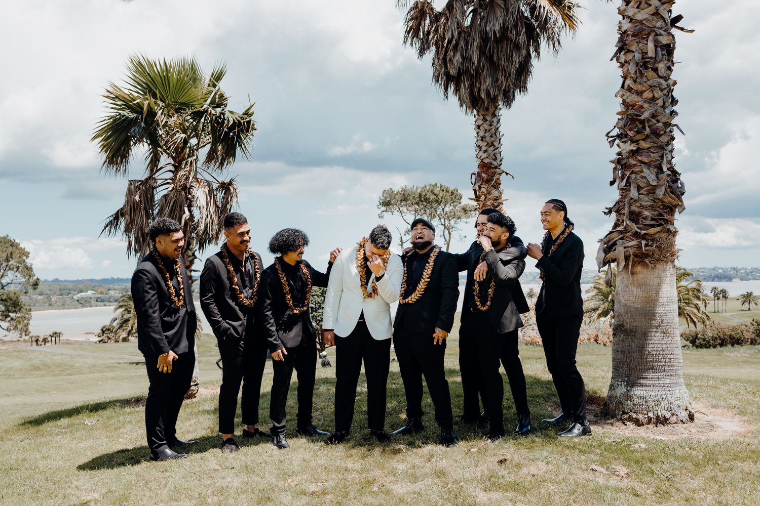 Wedding party in formal attire gathered under palm trees for group photo.