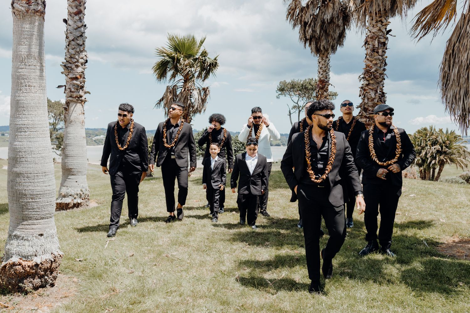 Groomsmen in matching black suits walking together outdoors with palm trees in background.