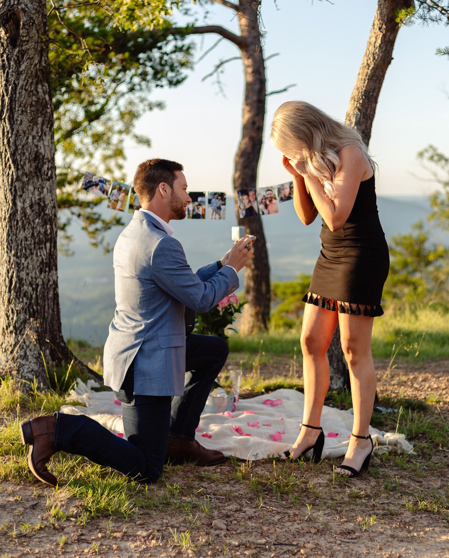 A man wearing a blue suit proposes to a woman wearing a black dress at sunset.