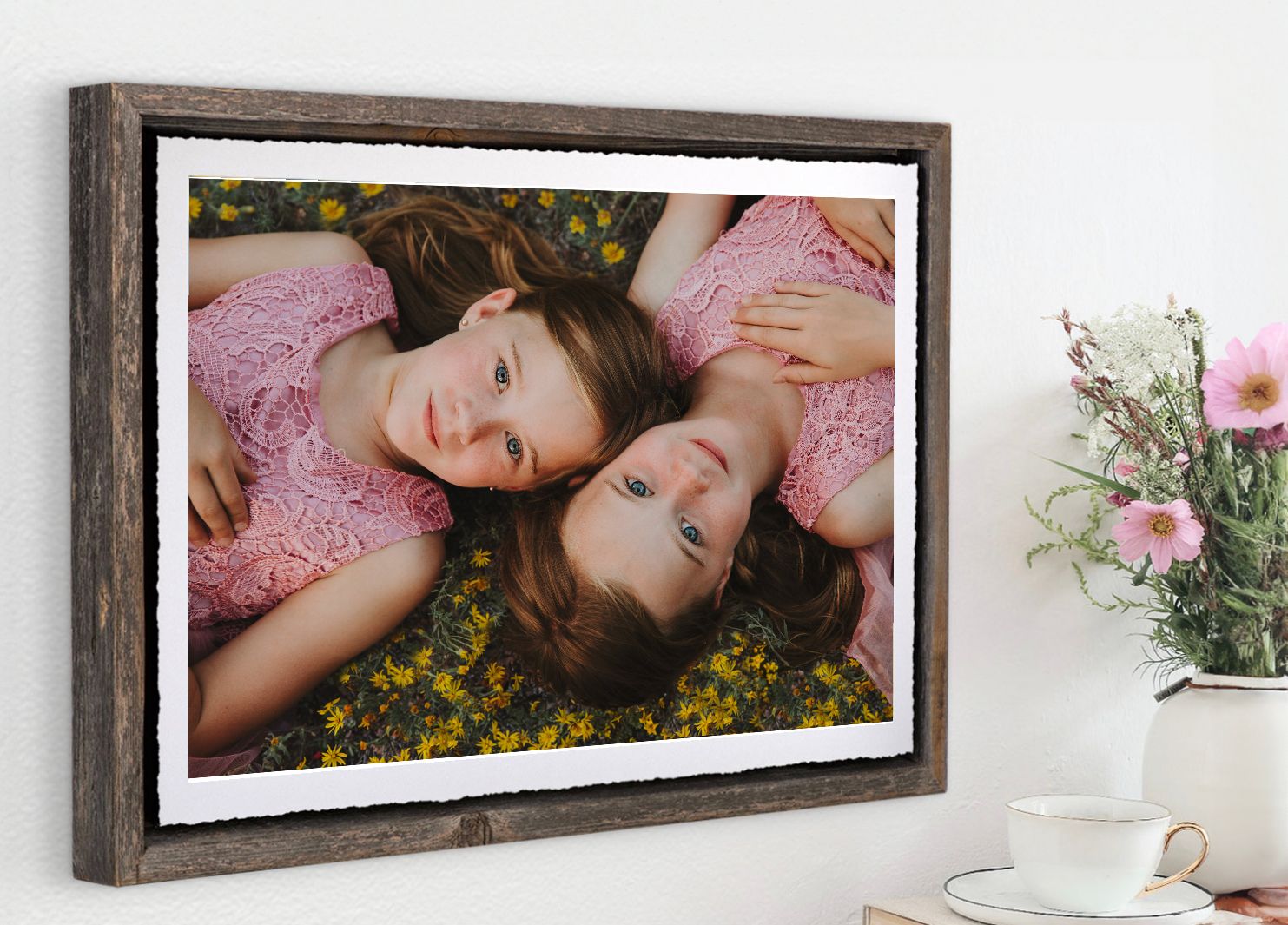 A framed photograph of two people lying in yellow flowers wearing pink attire displayed next to a flower vase.