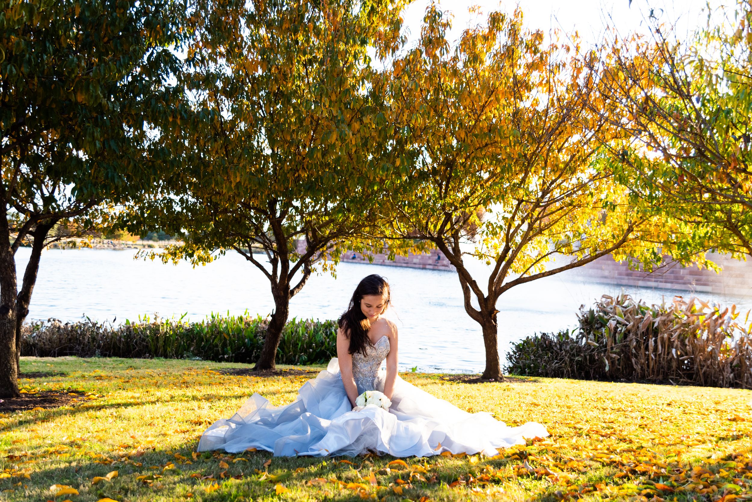 Bride in white dress sits on grass with golden sunlight filtering through trees.