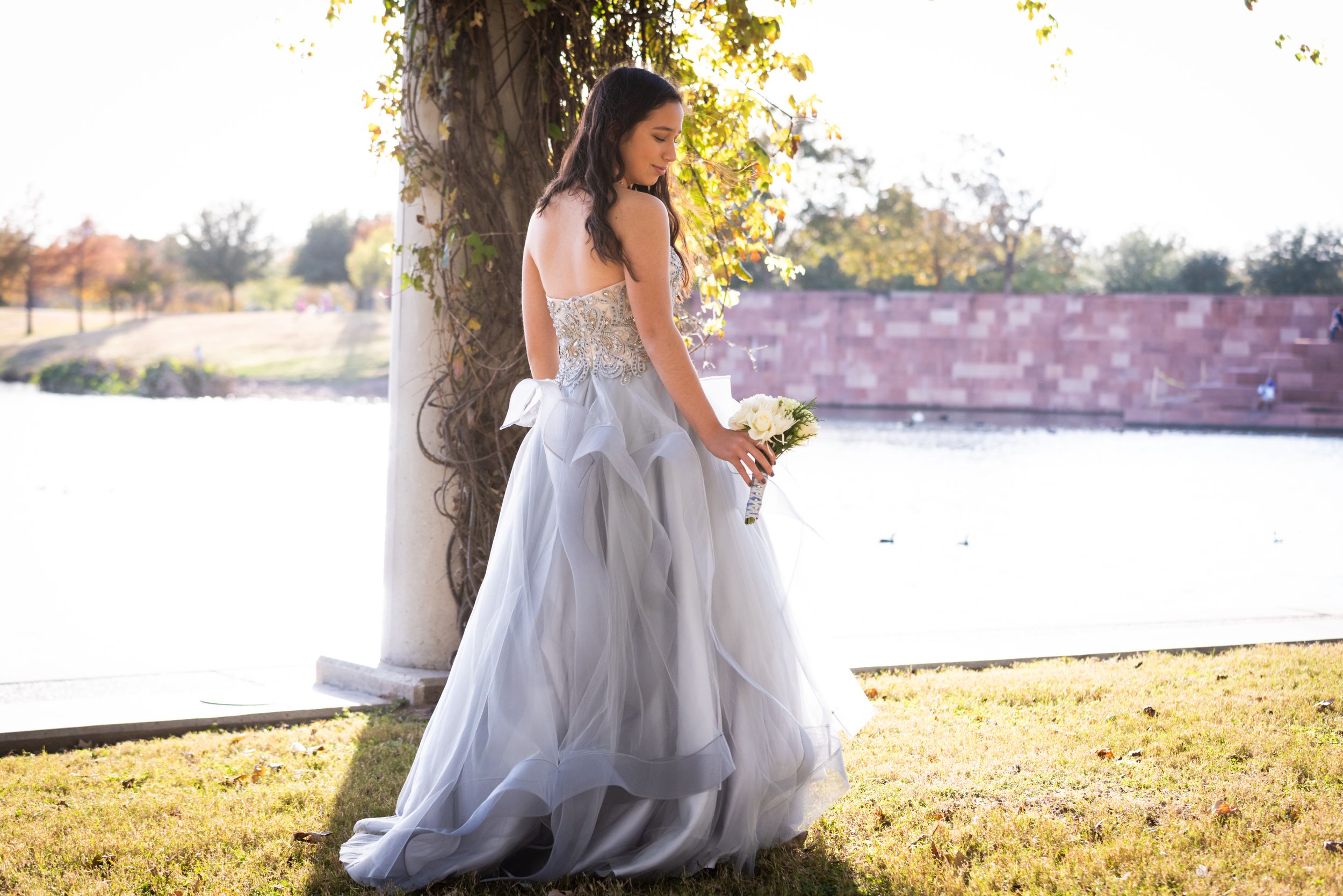 Bride in strapless gown poses by lake during golden hour with bouquet.
