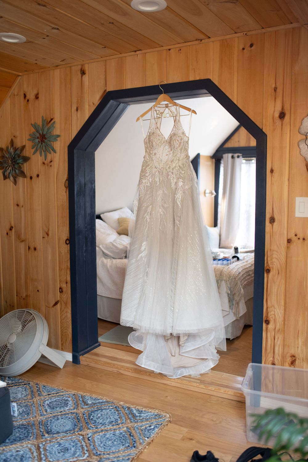 A beaded wedding dress hangs in a hexagonal mirror frame against wooden paneled walls.