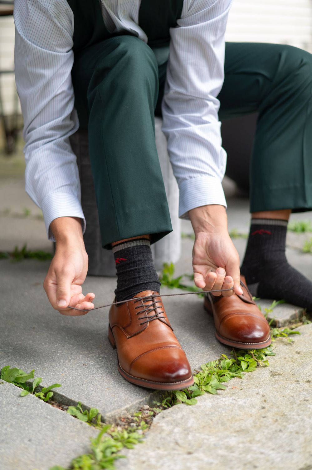 Brown leather dress shoes being laced up with green formal pants and white dress shirt.