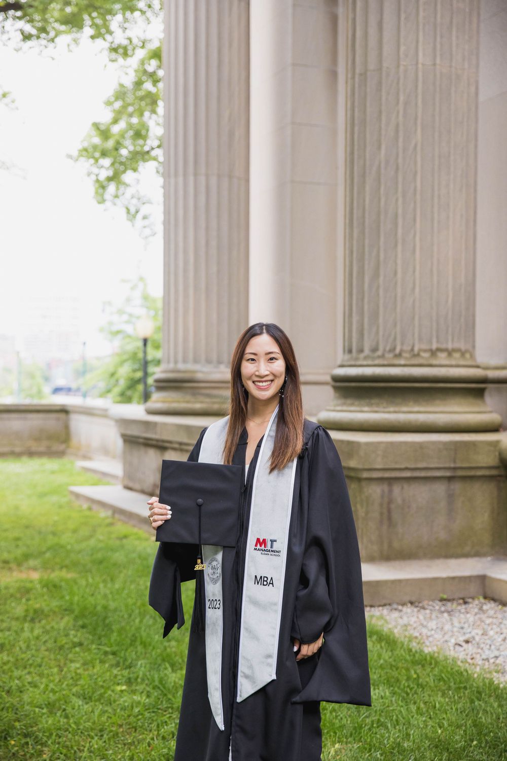 Graduate holding diploma and wearing academic regalia poses next to MIT's classical stone columns.