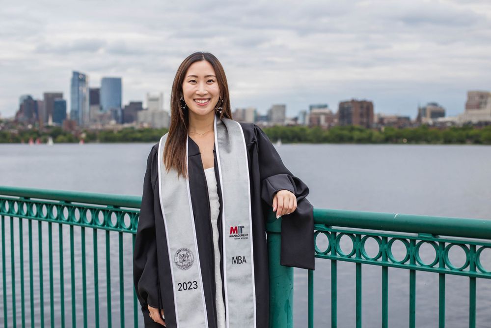Graduate in cap and gown poses on bridge with Boston skyline in background.