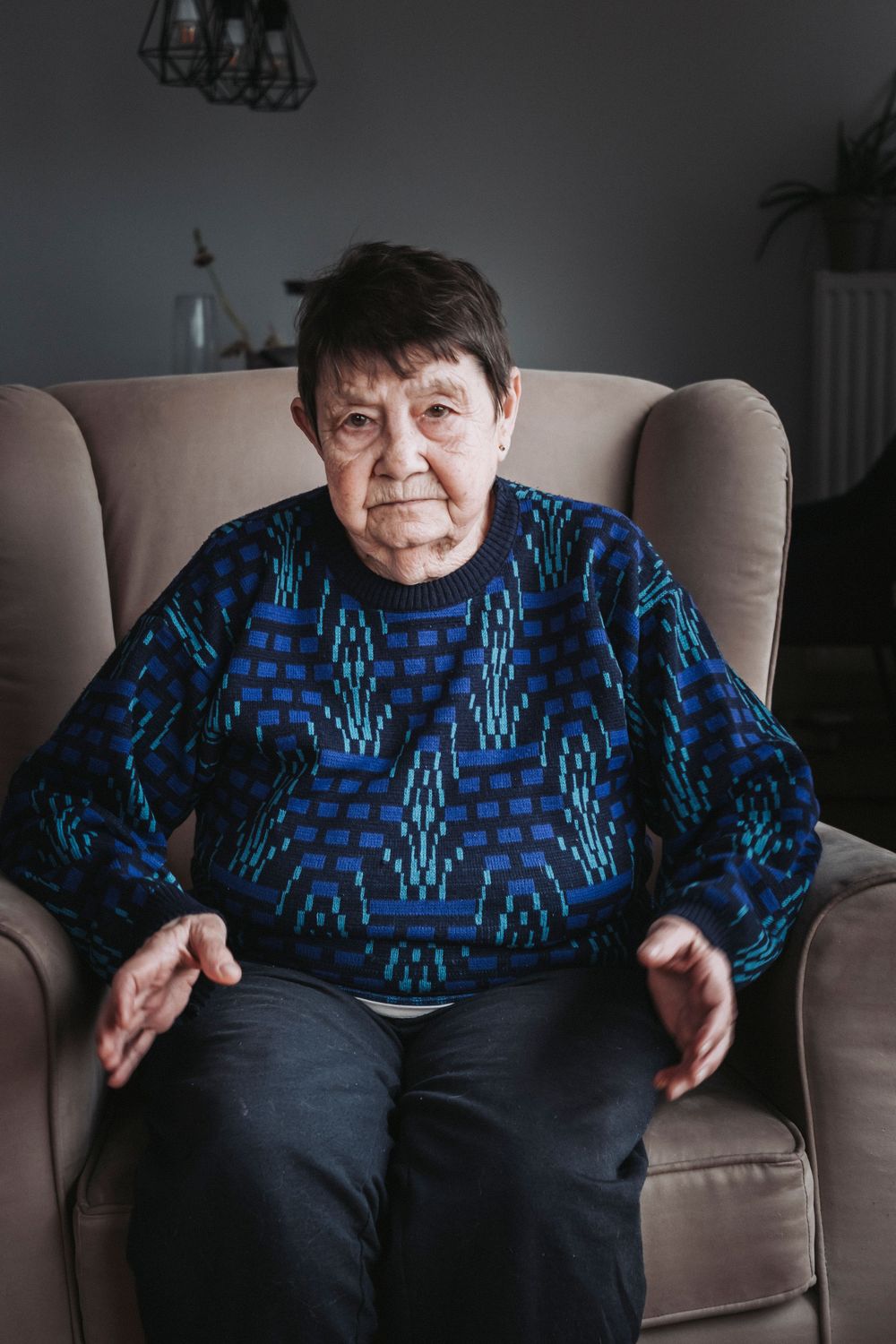Senior woman in blue patterned sweater sits in beige armchair against neutral background.