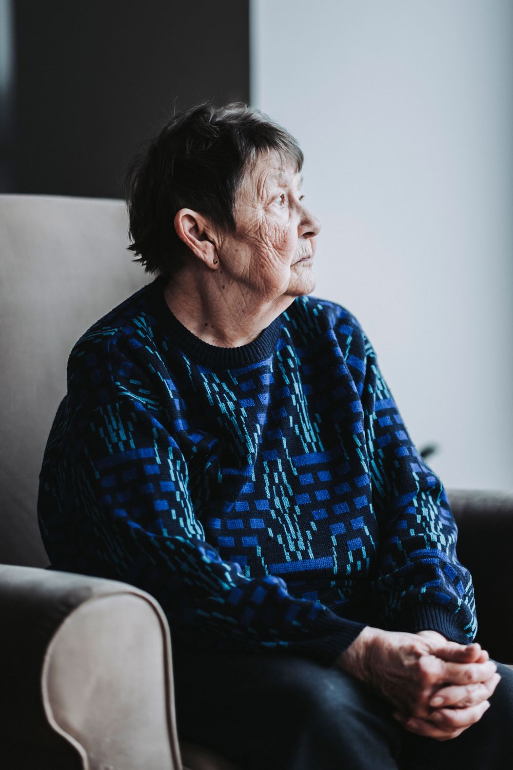Profile view of elderly person in blue geometric sweater sitting in armchair near window.