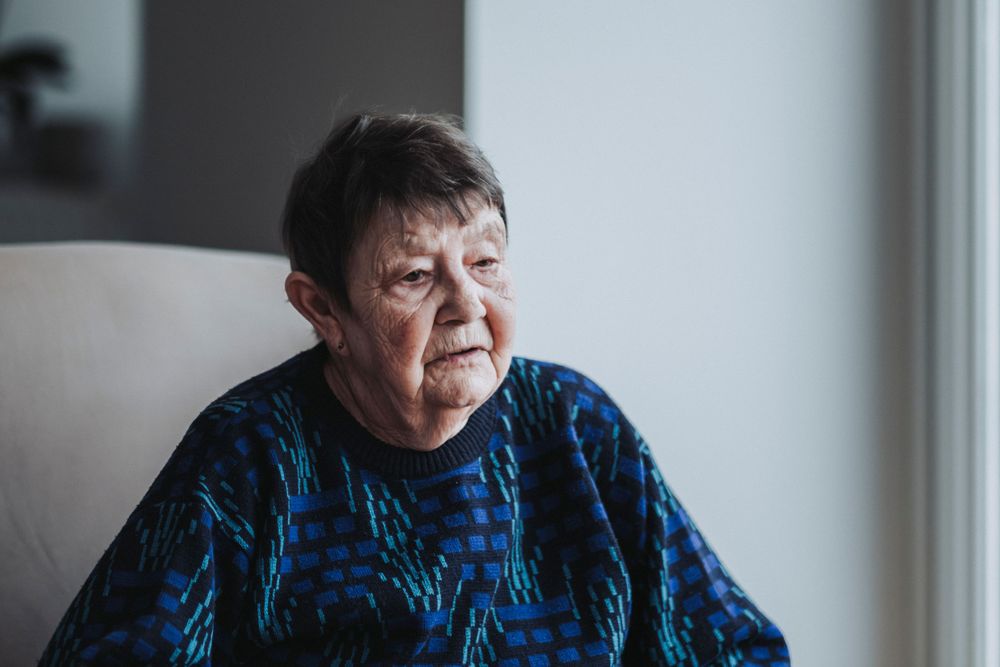 Senior in blue patterned sweater gazing thoughtfully out window while seated in armchair.