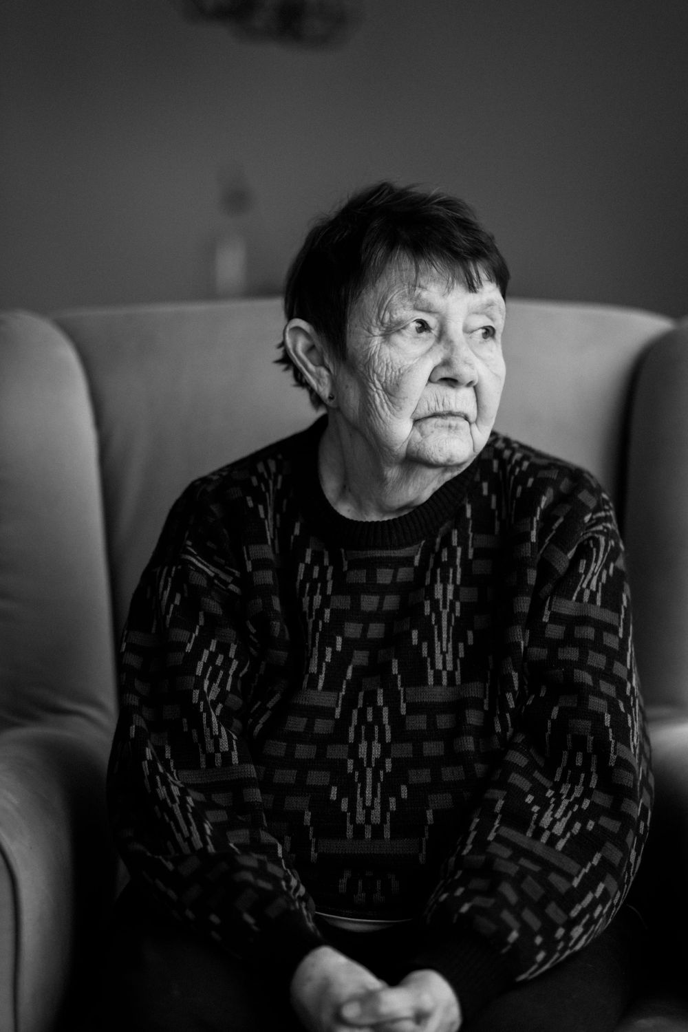 Portrait of elderly person in patterned sweater seated in armchair, captured in monochrome.