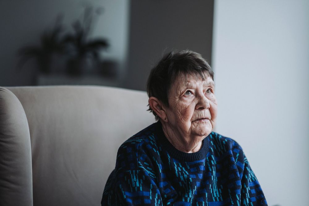 Senior wearing vibrant blue patterned sweater seated in beige armchair by window.
