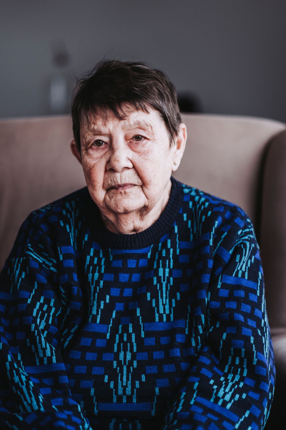 Senior wearing a geometric blue sweater sitting in a beige armchair by natural light.