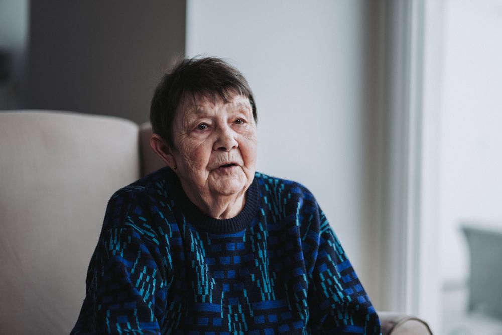 Elder in blue geometric sweater sitting in armchair with natural window lighting.