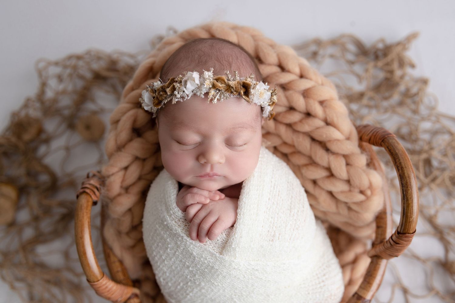 Sleeping newborn wrapped in white swaddle with floral crown lying in wooden basket on chunky knit brown blanket.