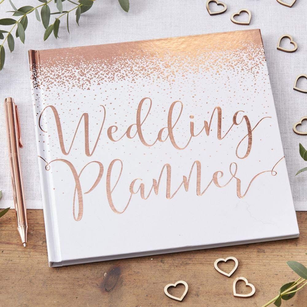 A rose gold wedding planner book with wooden hearts and eucalyptus leaves on white surface.