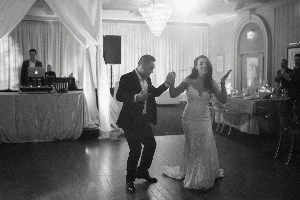 A couple dances enthusiastically in an elegant ballroom with white draping and a chandelier overhead.