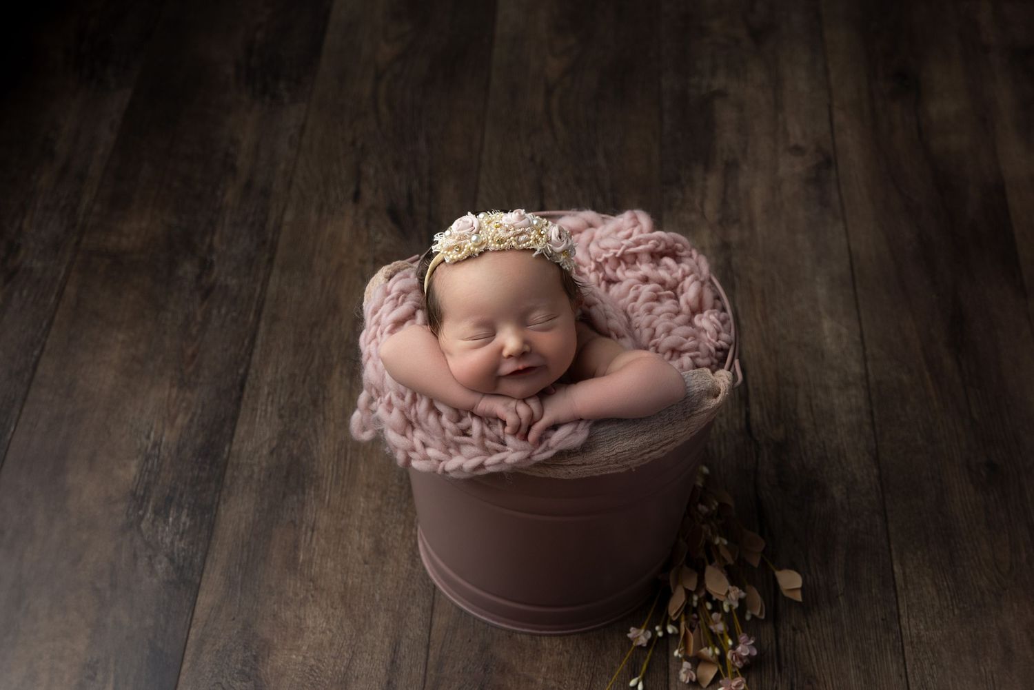 Sleeping newborn in pink blanket nestled in brown bucket on wooden floor with rustic backdrop.