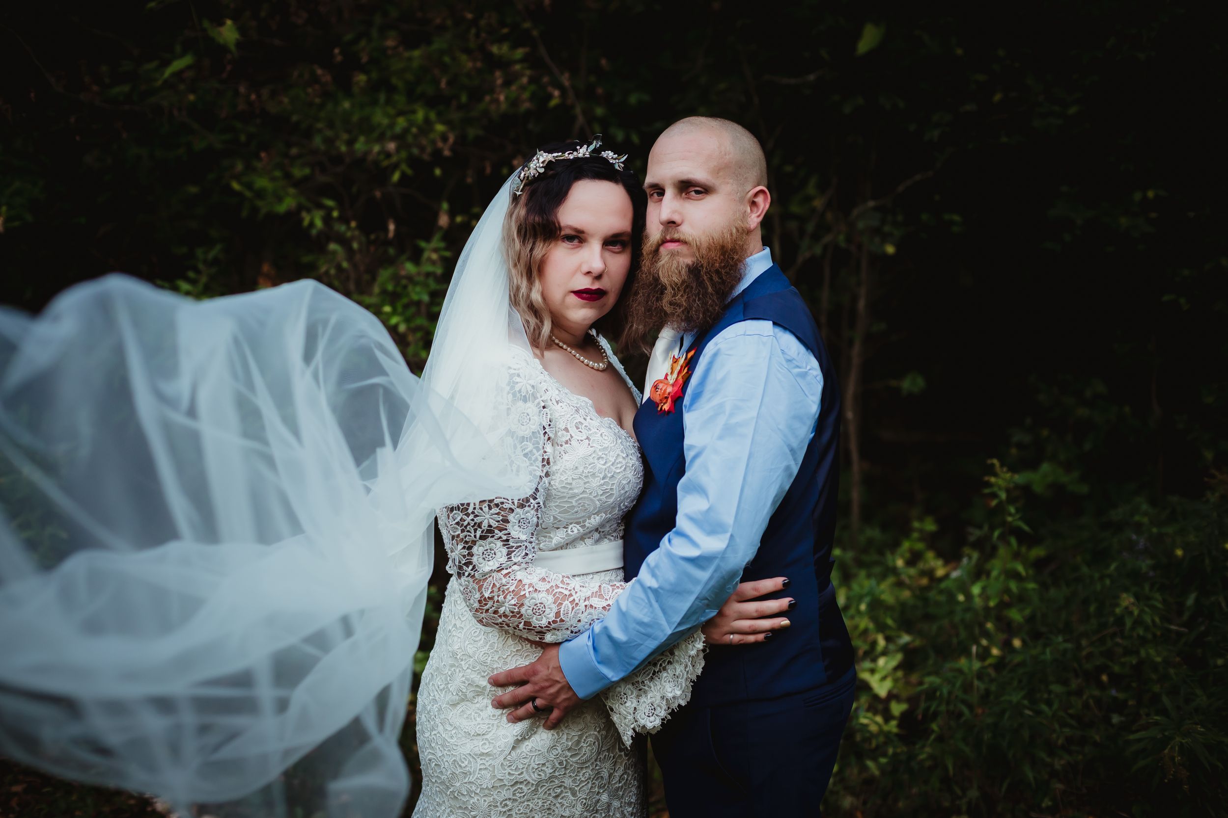 Woman and man in wedding attire looking seriously at the camera. Her long veil is held toward the camera.