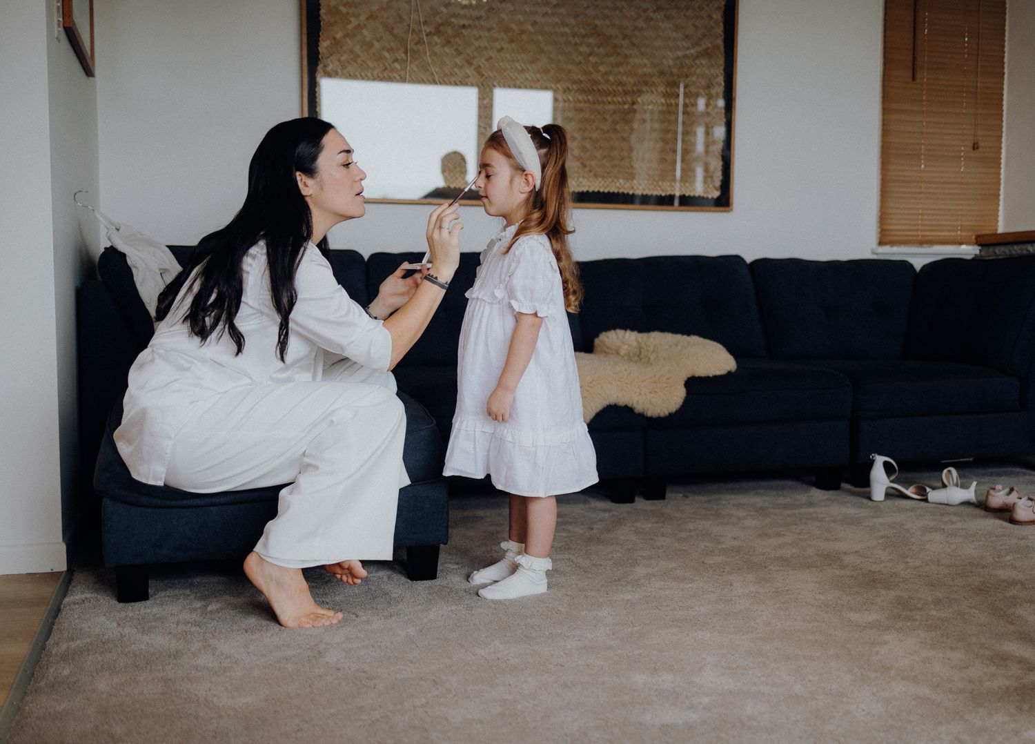 A woman and child in white outfits share a playful moment in front of a navy blue couch.