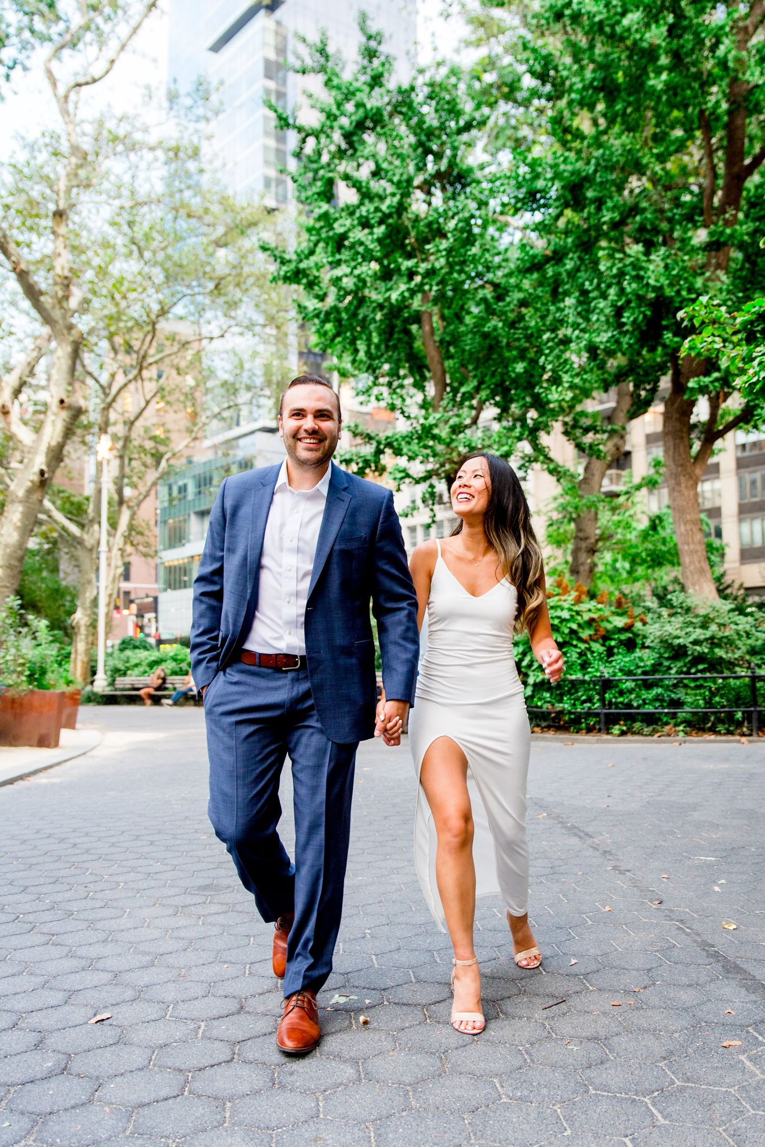 young engaged couple in NYC walking through Madison Square Park in white dress and navy suit