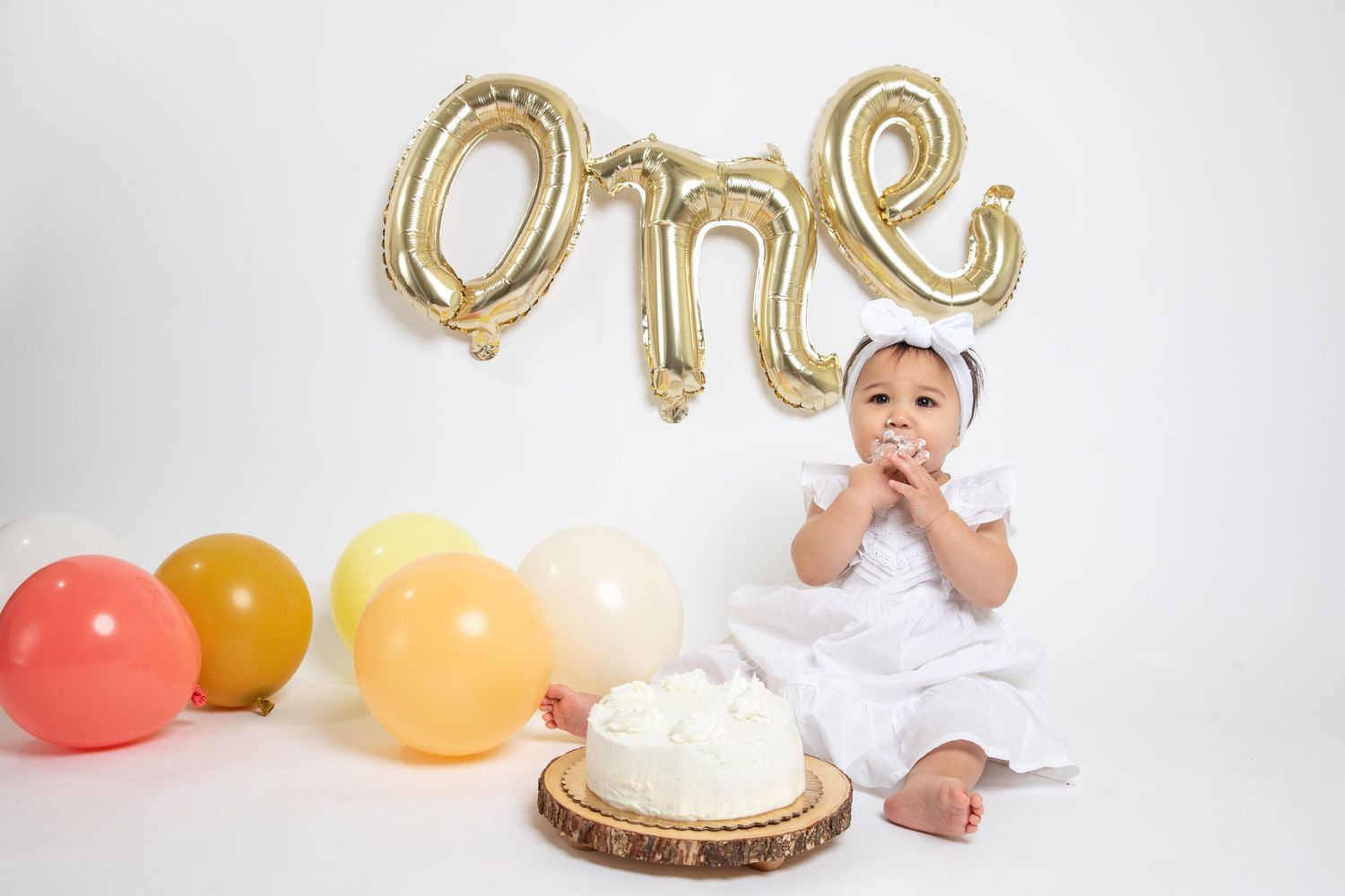Baby in white outfit sitting by gold ONE balloon letters and colorful balloons with birthday cake.