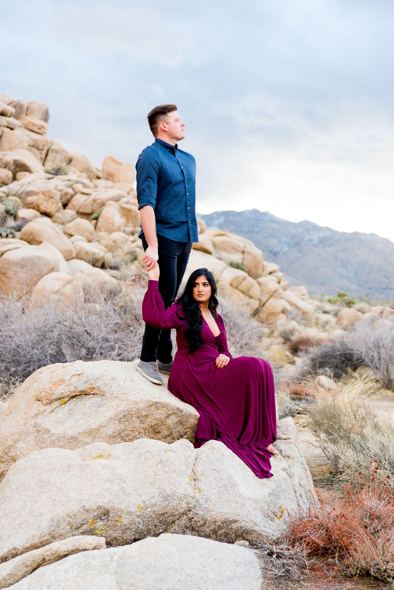 man in blue button down stands on rocks at Joshua Tree National Park and holds hand of woman in red dress who is sitting