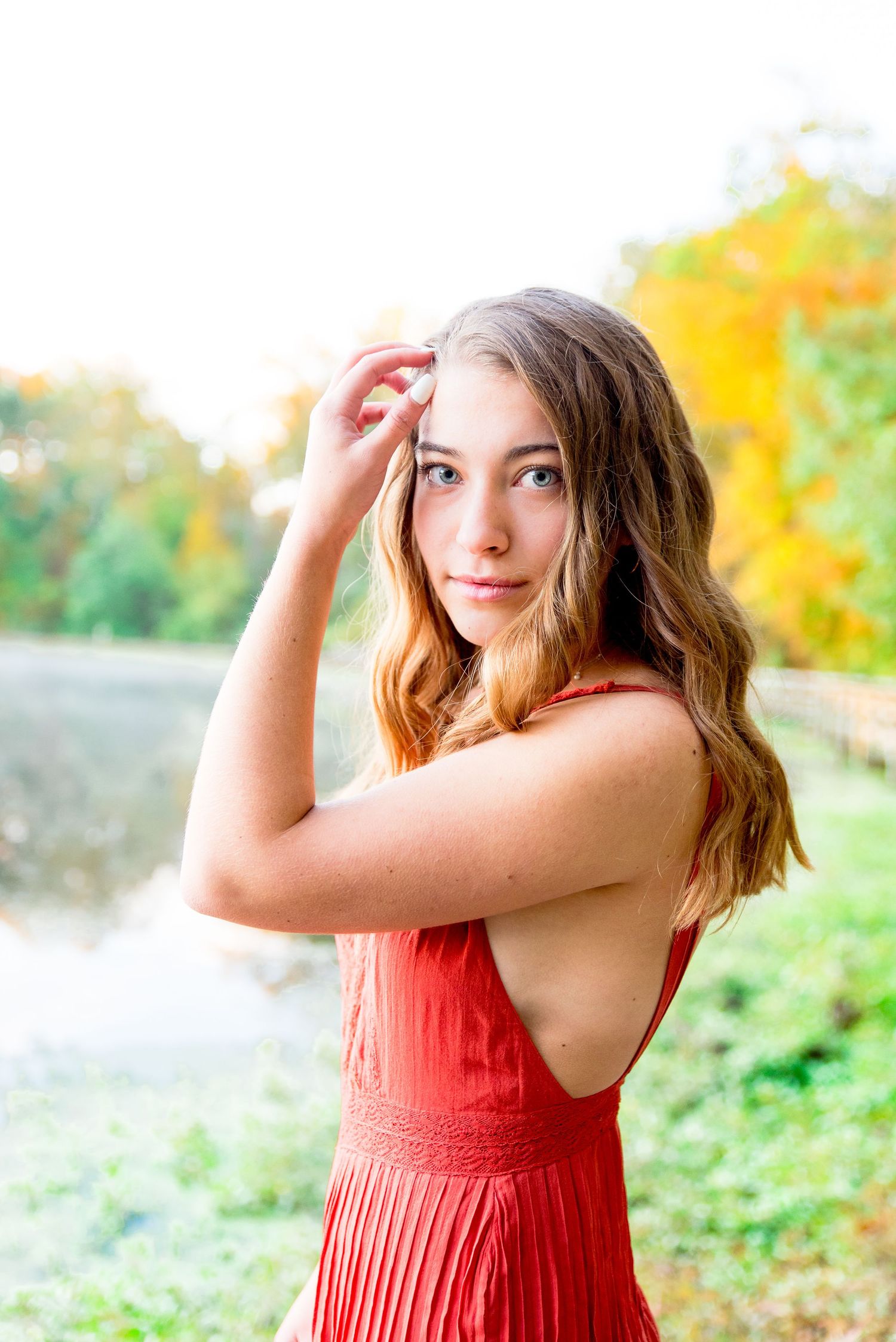 young woman with blue eyes puts hand on face in front of fall trees for senior pictures in Chicago