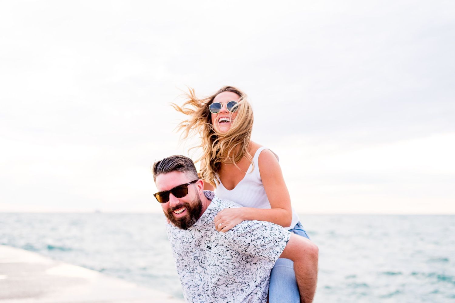 woman in white tank top and sunglasses piggyback riding on her dark haired fiance in sunglasses at North Ave Beach