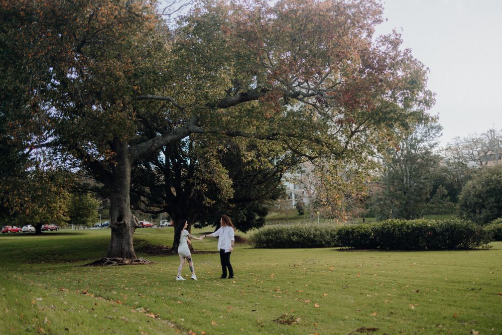 Two figures embrace while standing on a green lawn with fall foliage in the background.