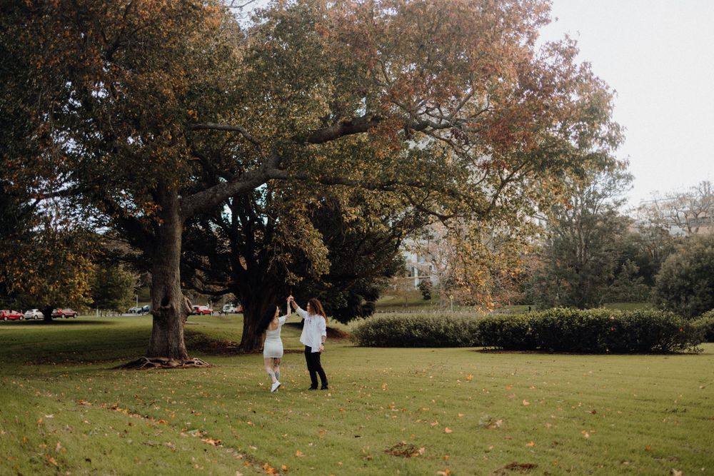 Two people share an intimate moment while standing under large trees in a park setting.