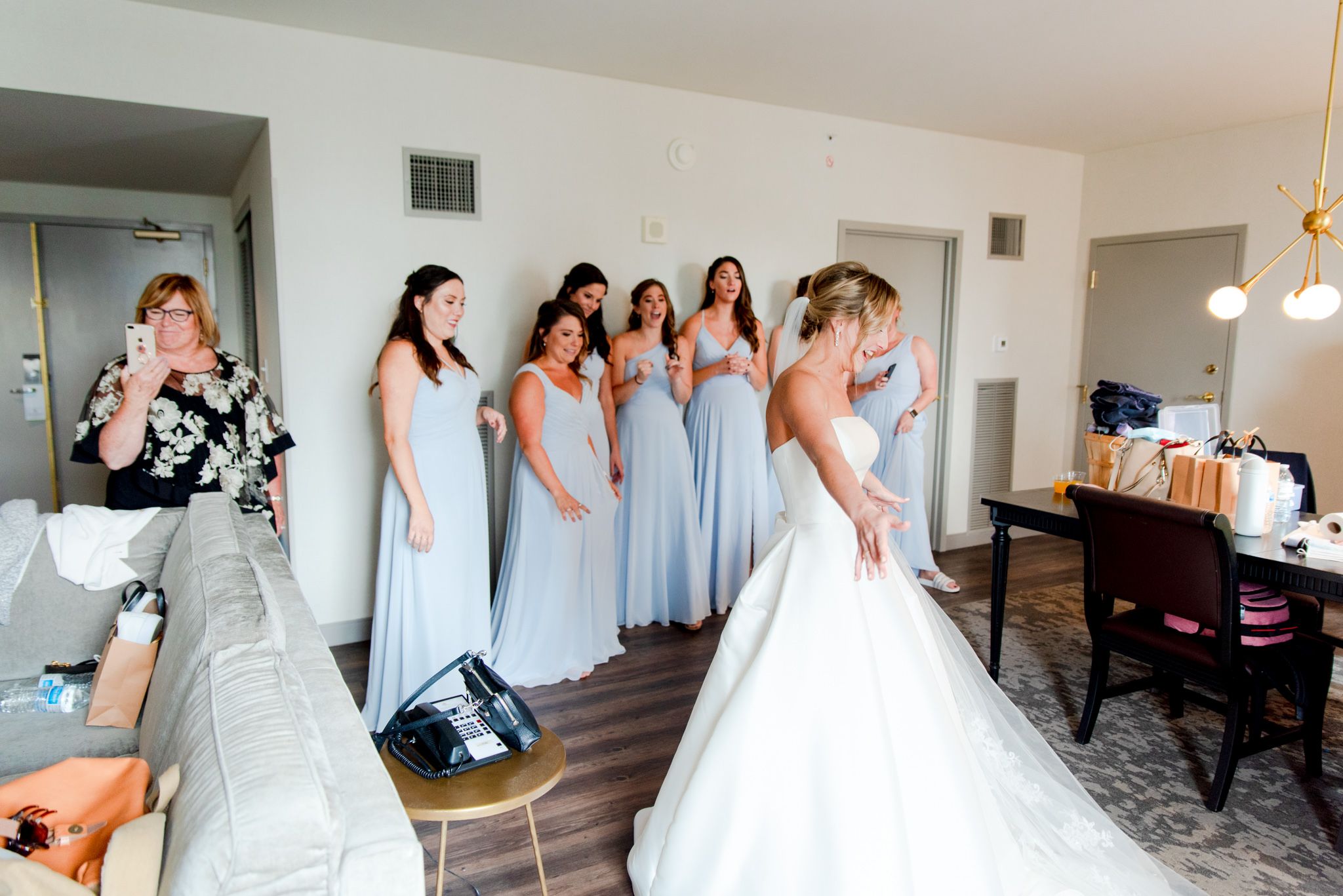 7 bridesmaids in light blue dresses smiling and clapping after seeing the bride, while mom smiles and holds up phone