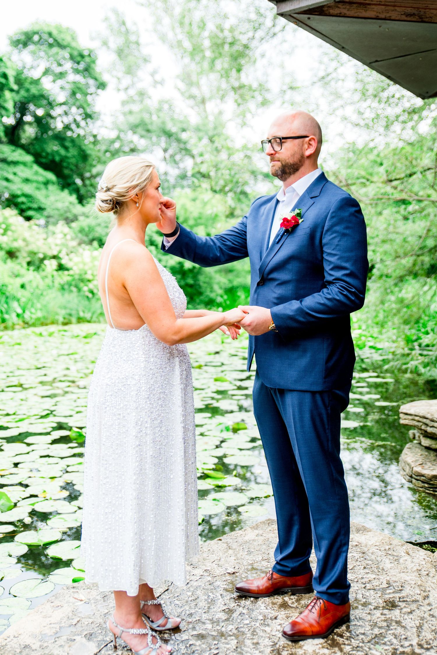 man in navy suit drying wife's tears on a rock at Alfred Caldwell Lily Pool for Chicago summer elopement