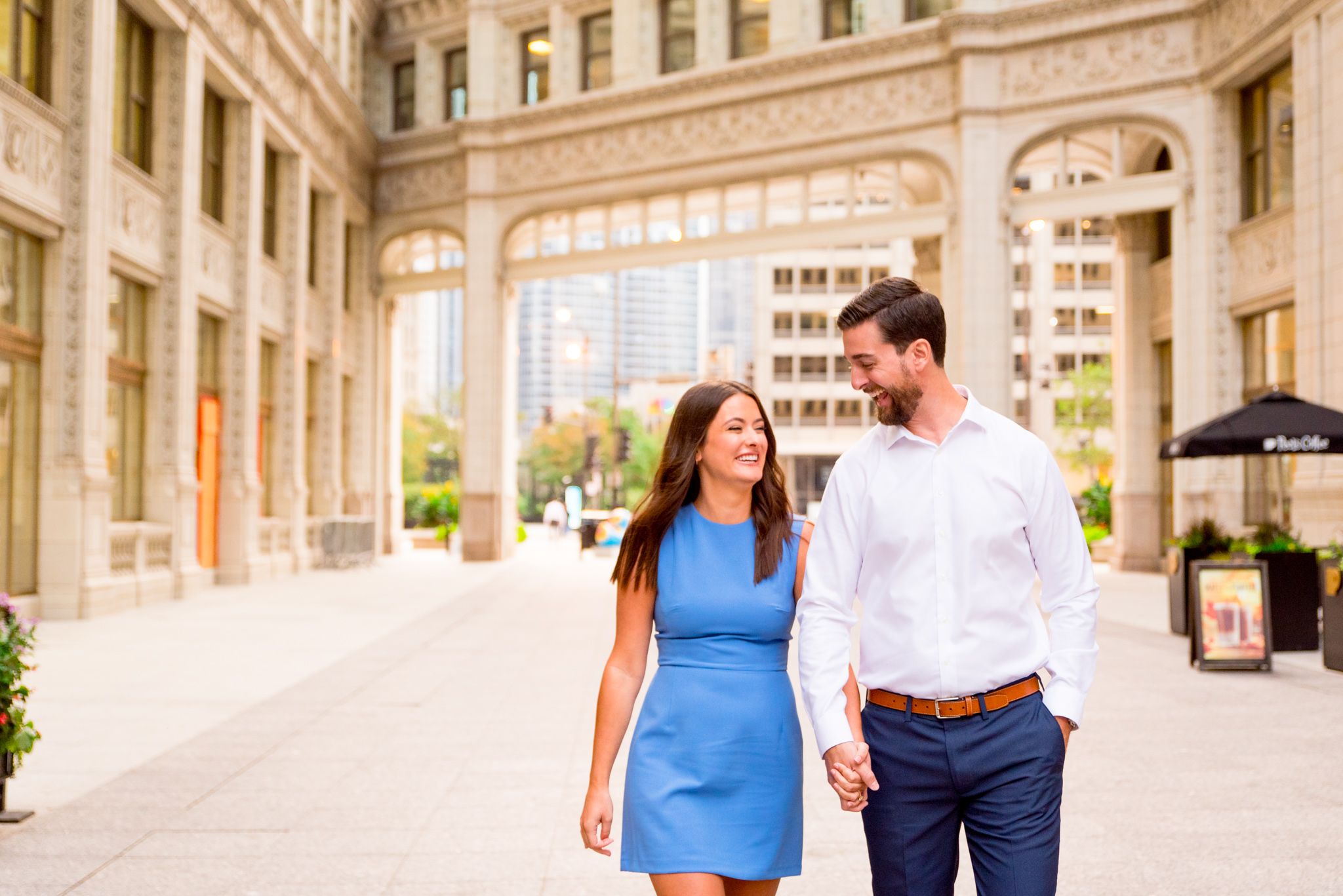woman with brown hair in a light blue dress holding hands with fiance in front of the wrigley building in chicago