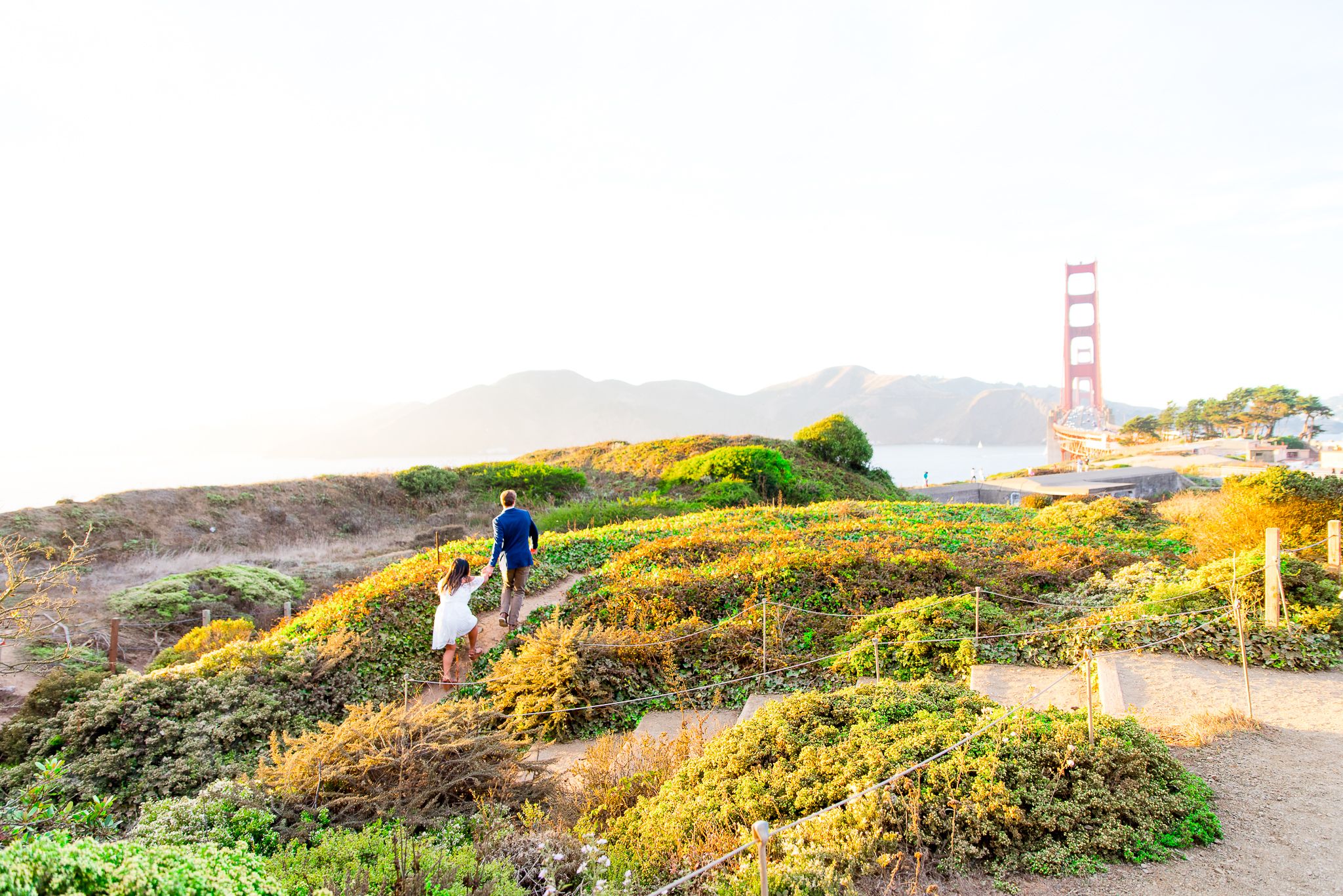 man in navy suit leading woman in white dress through hills near Golden Gate Bridge engagement pictures