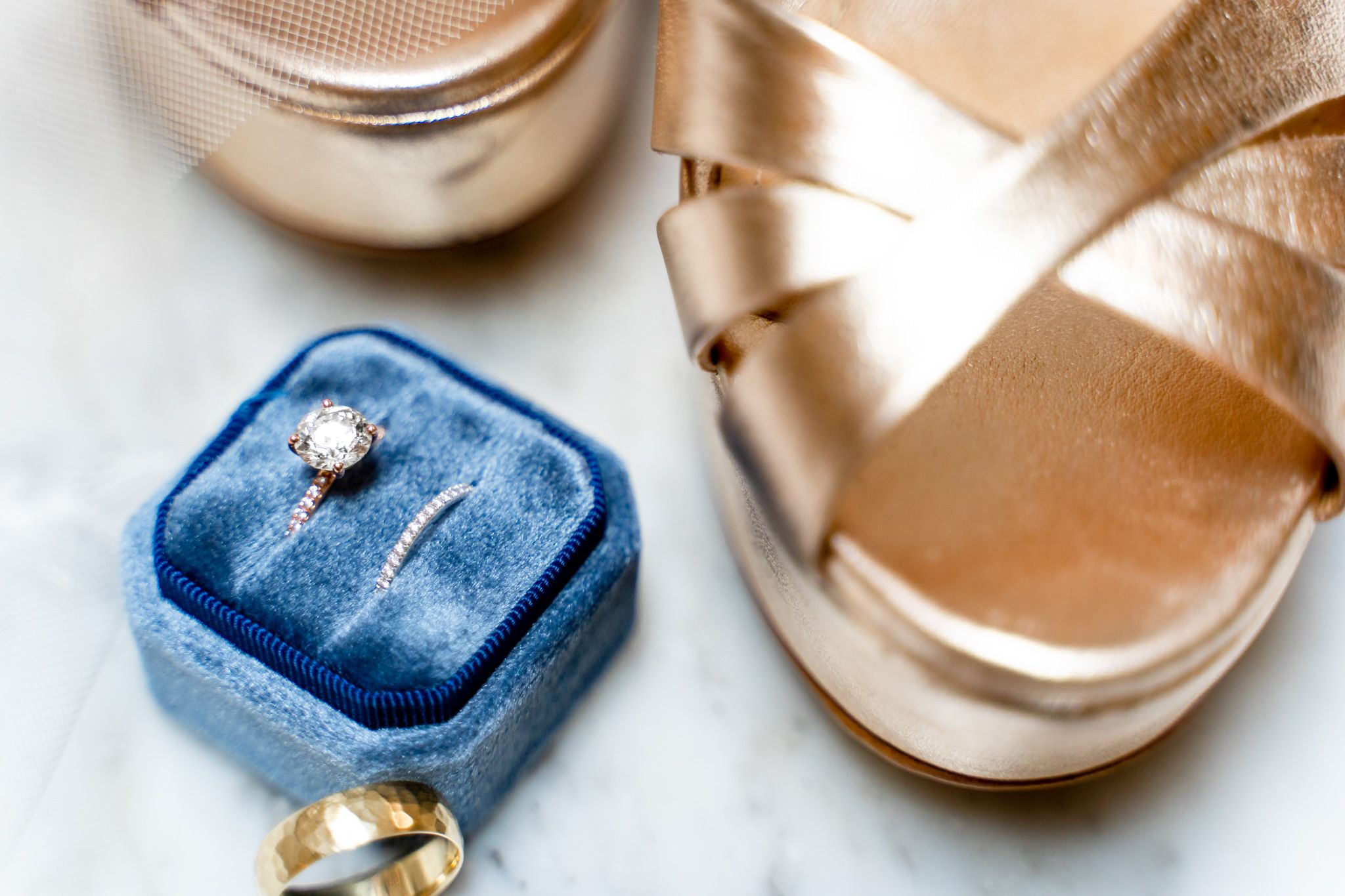 round diamond engagement ring with wedding bands in blue velvet ring box on lace wedding dress and gold wedding shoes