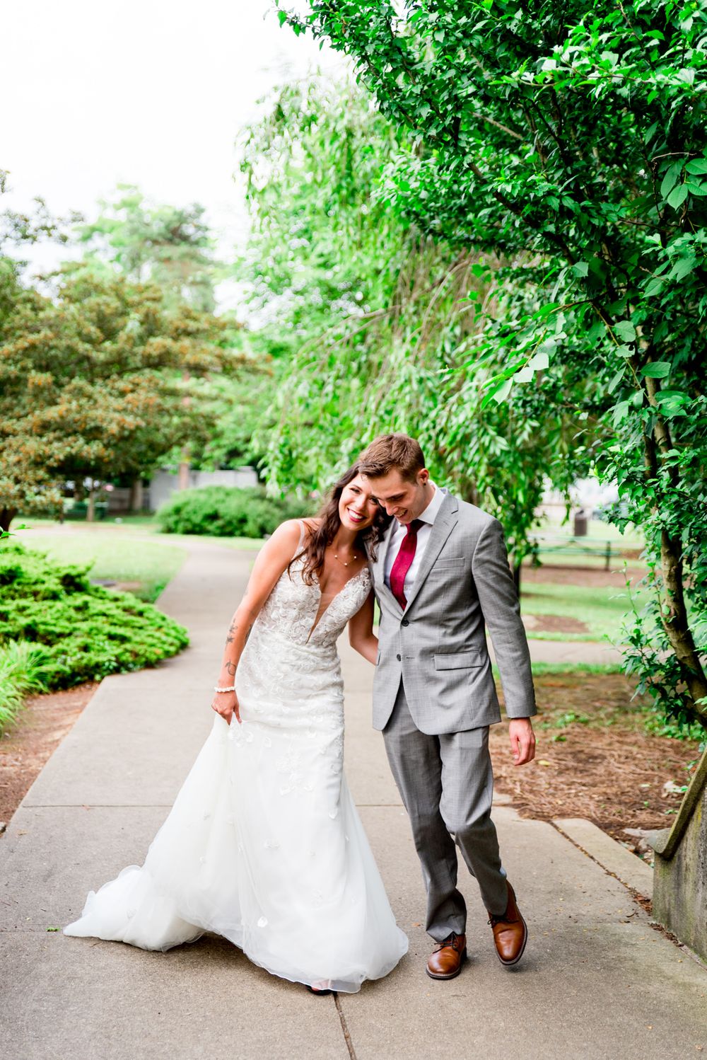 bride in Serafina gown walks and lays on chest of groom in gray suit and red tie at Eden Park wedding