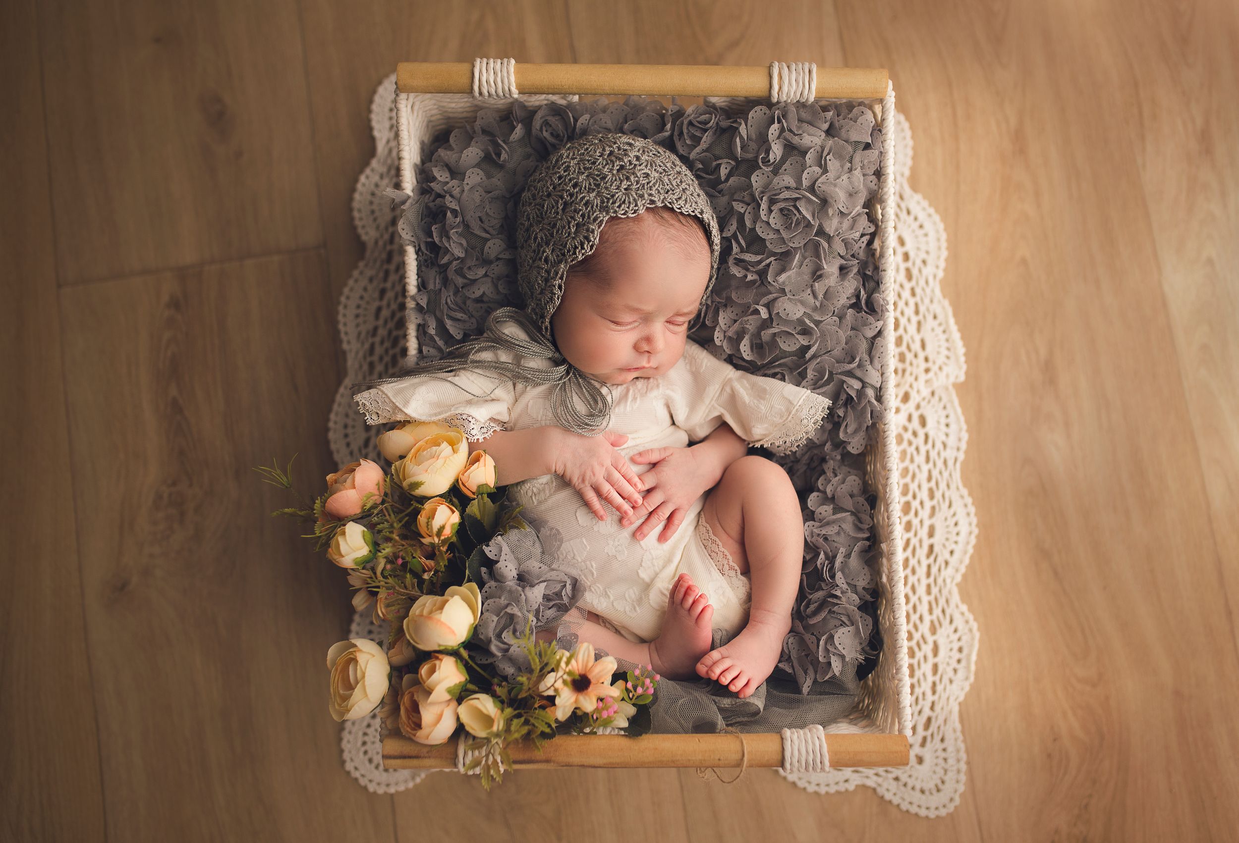 newborn baby girl wears a gray bonnet laying in square basket with flowers