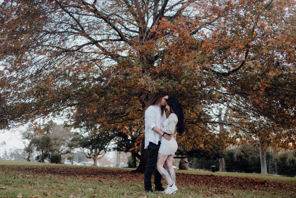 A couple shares a romantic embrace under a large tree with autumn leaves.