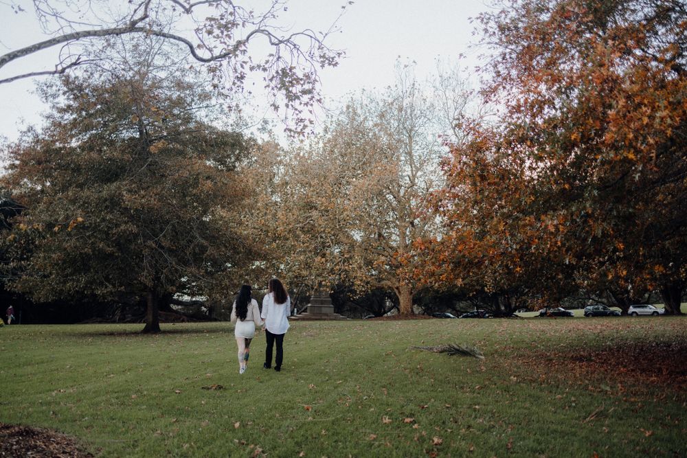A couple strolls through a park with autumn trees and fallen leaves surrounding them.