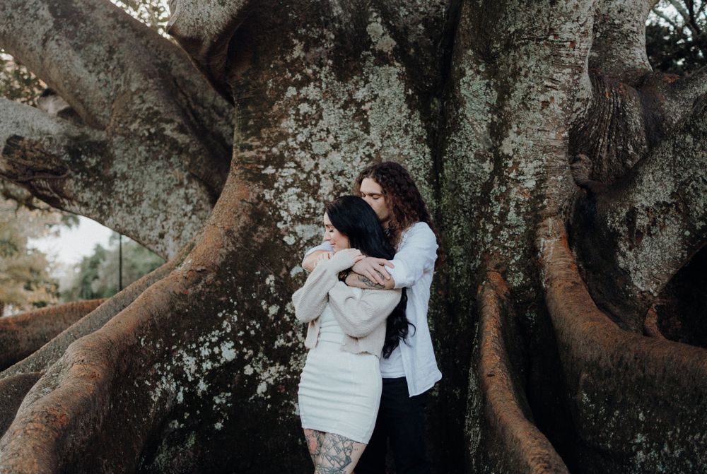 Two people embrace near the twisted roots of a large, ancient tree.