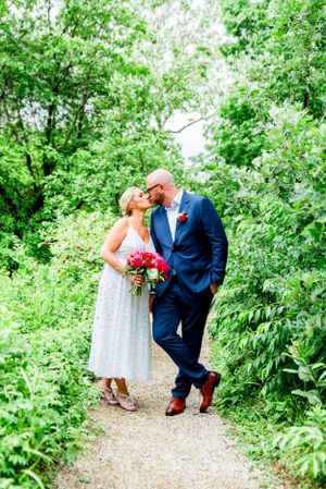 man in navy suit kissing blonde woman in silver dress with red bouquet at Alfred Caldwell Lily Pool in Lincoln Park