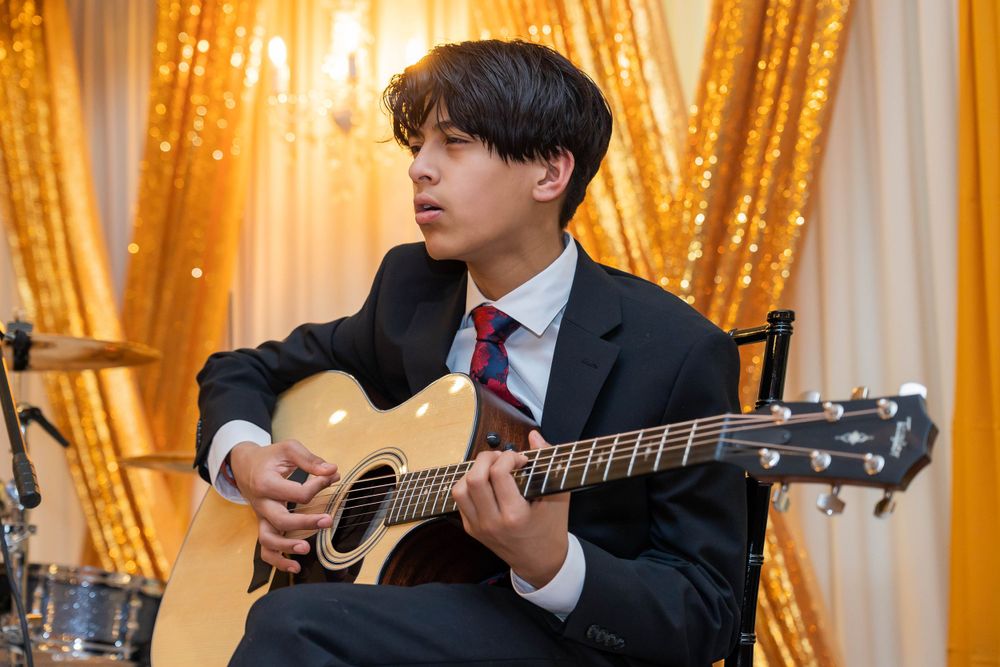 Musician in formal suit plays acoustic guitar during performance with golden backdrop.