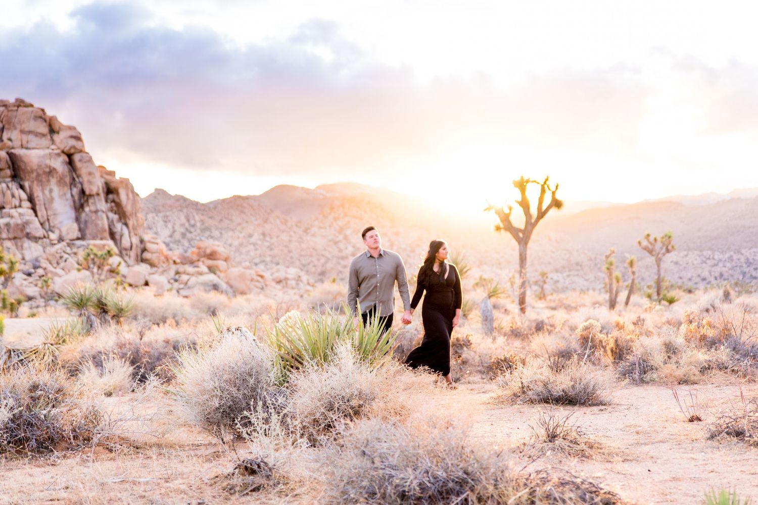 woman in black dress holds man's hand and leads him through Joshua Tree National Park at sunset for engagement pictures