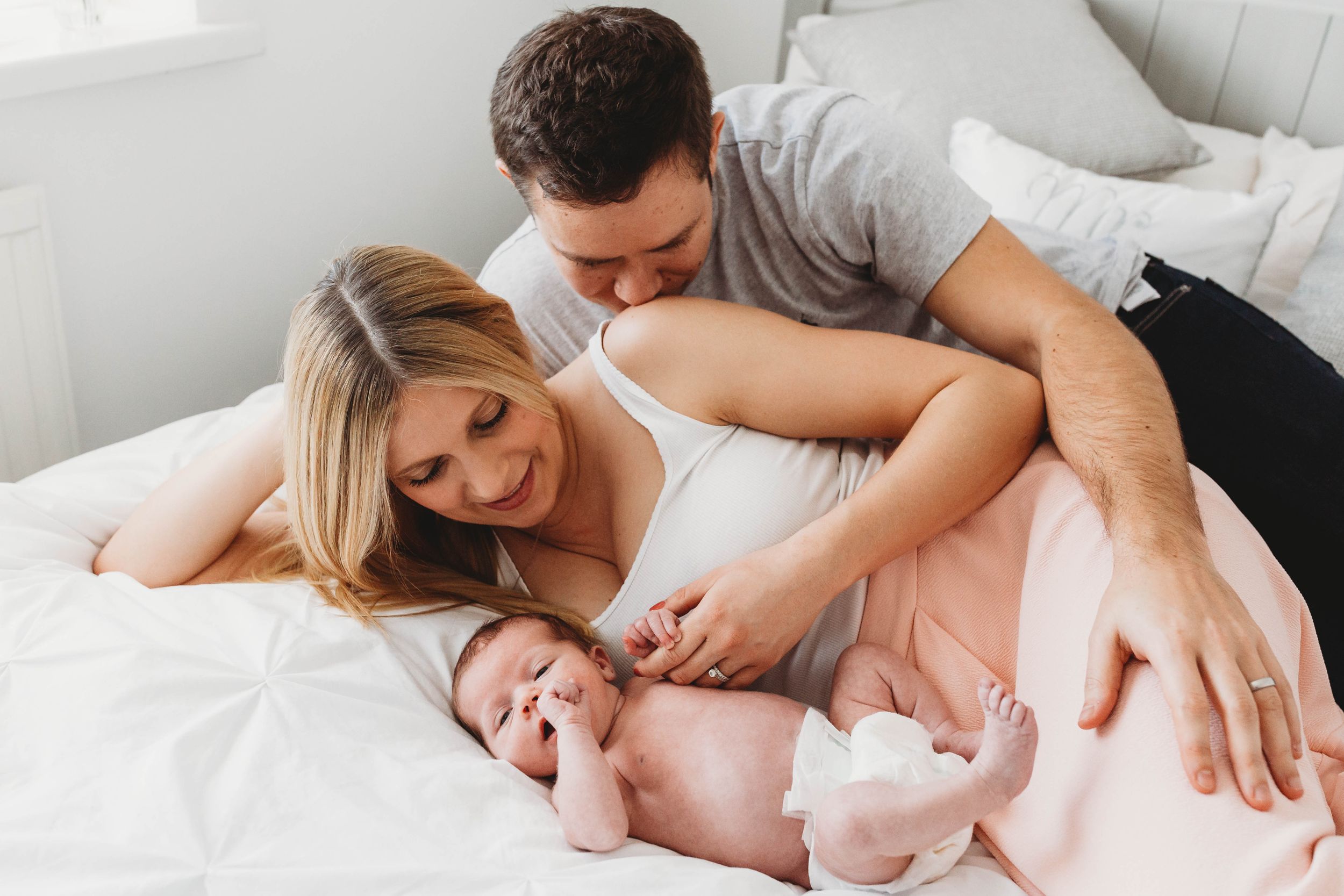 two week old baby girl on bed with mum dad kissing woman shoulder