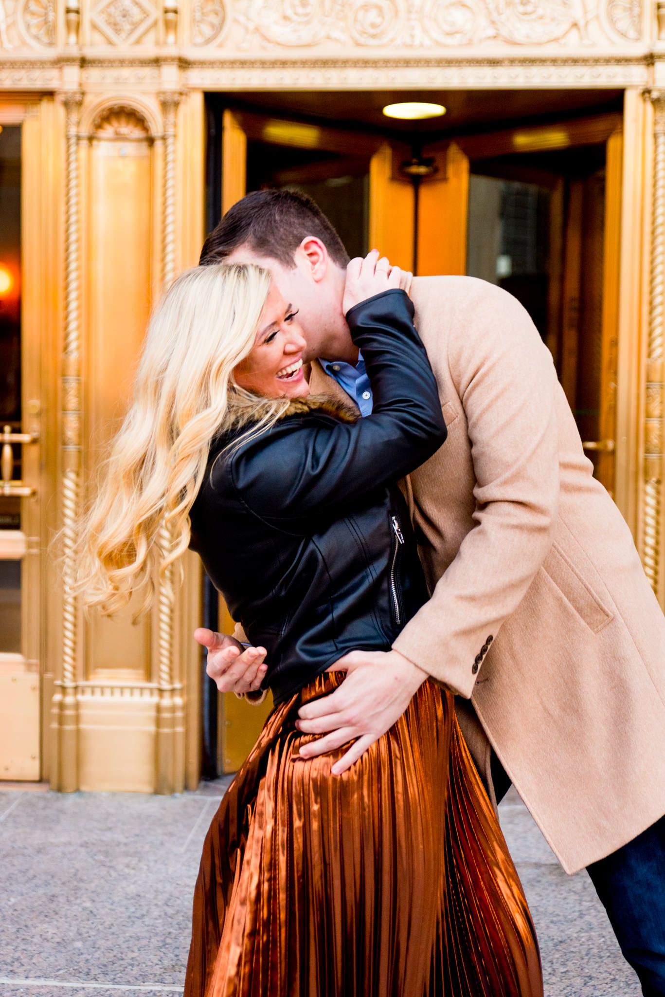 man in tan coat laughing with woman in black coat and copper skirt in front of gold doors at Wrigley Building engagement