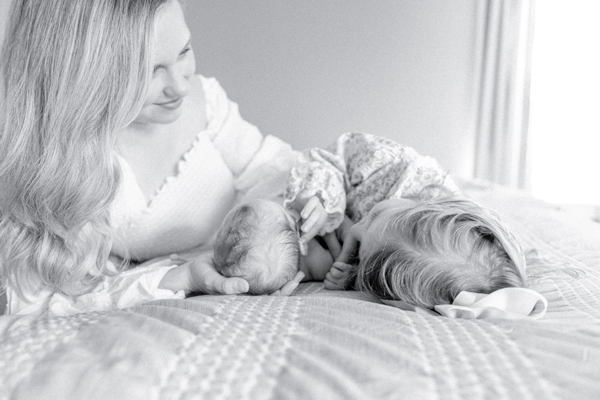 Mother snuggles on a bed with her newborn son and toddler daughter as they lay head first.