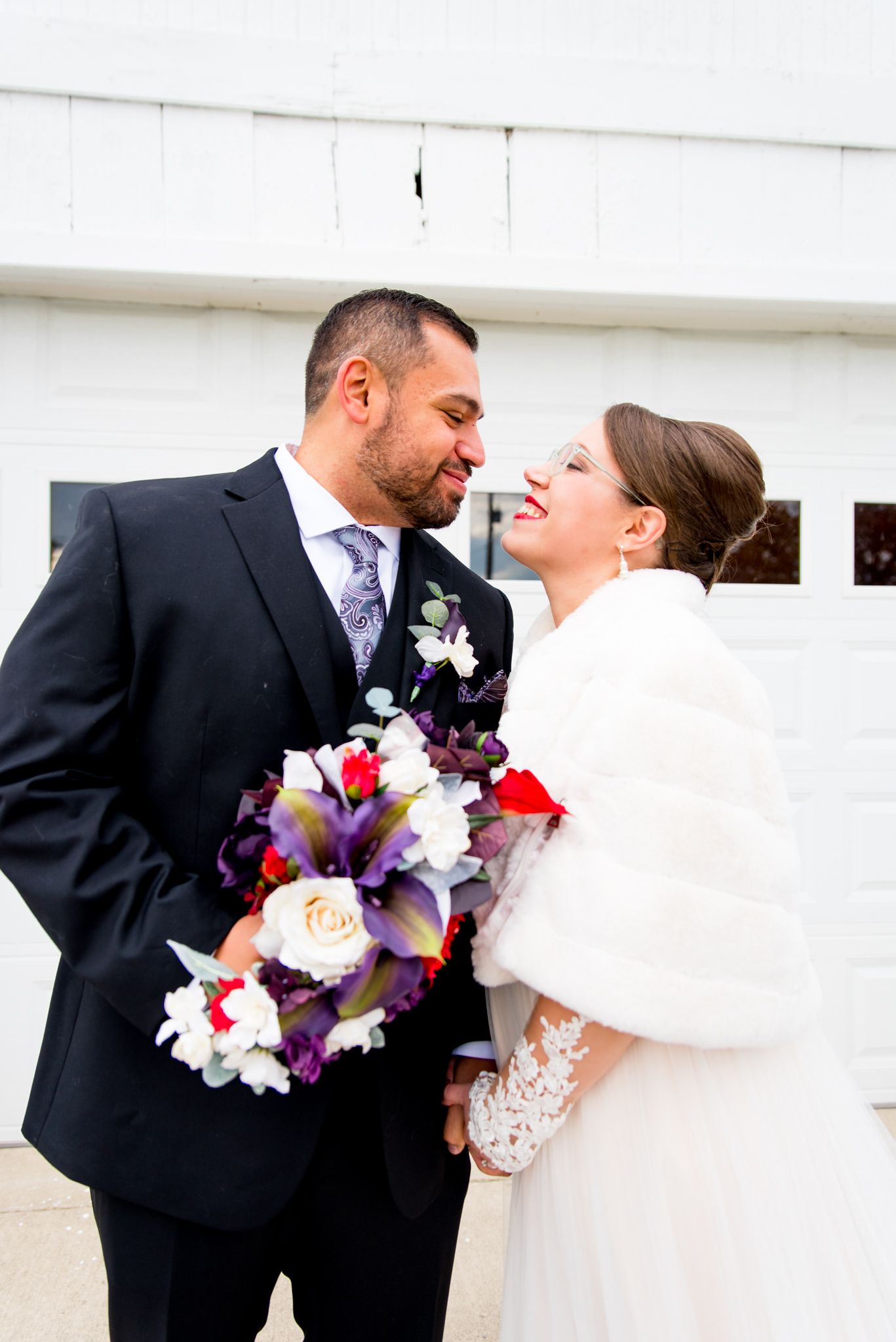 bride in red lipstick smiling at groom in purple tie while they hold purple and red wedding bouquet